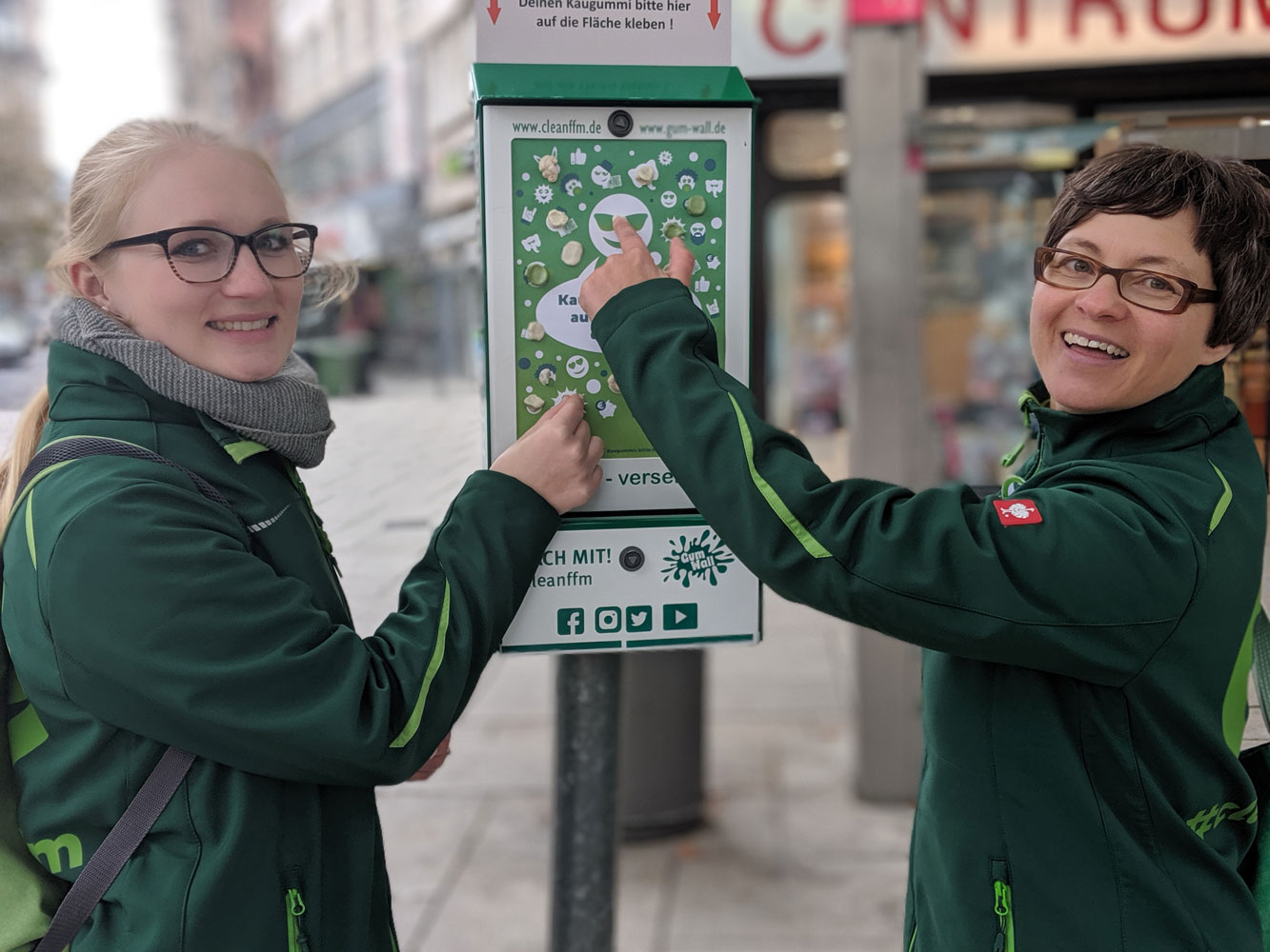 Foto: Zwei Mitarbeitende zeigen auf eine cleanffm Gum Wall 